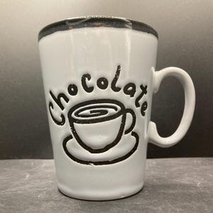 Op de afbeelding: Een witte keramische mok met een zwarte rand en handvat. Het woord "chocolate" is in zwarte letters gegraveerd boven een zwarte afbeelding van een kopje koffie op een schotel. De mok is taps toelopend en heeft een gladde afwerking.