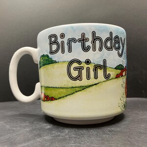 以下が含まれることがあります： 白いセラミックマグカップ、ハンドル付き、黒い縁取り文字で「Birthday Girl」の文字が特徴です。マグカップには、緑の畑、赤い納屋、青い空のある牧歌的なシーンがあります。