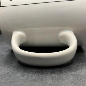 Puede incluir: Primer plano de una taza de cer&aacute;mica blanca con un asa curva. El asa es gruesa y redondeada, y la taza tiene un acabado brillante. La taza est&aacute; sobre una superficie gris.