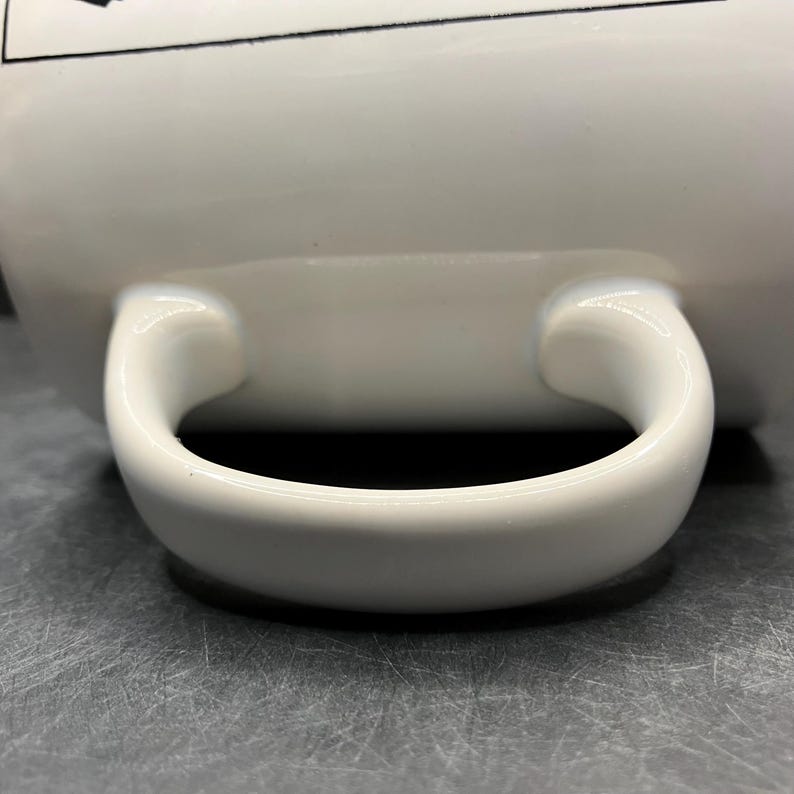 Puede incluir: Primer plano de una taza de cer&aacute;mica blanca con un asa curva. El asa es gruesa y redondeada, y la taza tiene un acabado brillante. La taza est&aacute; sobre una superficie gris.