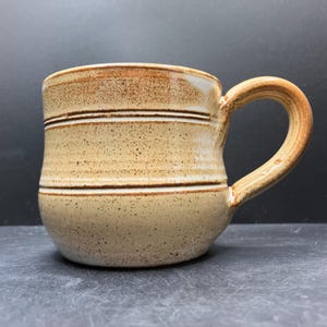 May include: A handmade ceramic mug with a speckled beige glaze and brown horizontal stripes. The mug has a rounded body, a sturdy handle, and a capacity suitable for coffee or tea. The stoneware mug is a rustic addition to any kitchen.