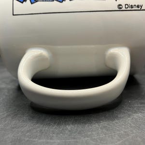 Puede incluir: Taza de cer&aacute;mica blanca con asa curva. La taza tiene un dise&ntilde;o en la parte superior con elementos azules y blancos. El logotipo de Disney es visible en el lado derecho de la taza.