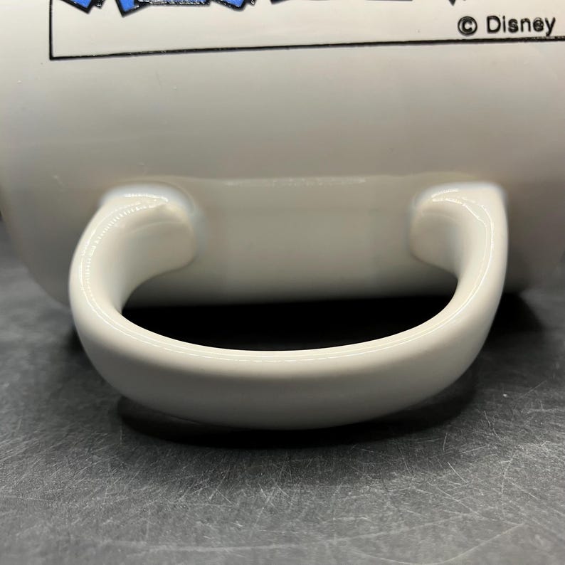 Puede incluir: Taza de cer&aacute;mica blanca con asa curva. La taza tiene un dise&ntilde;o en la parte superior con elementos azules y blancos. El logotipo de Disney es visible en el lado derecho de la taza.