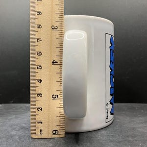 Puede incluir: Taza de cer&aacute;mica blanca con asa curva. La taza tiene un gr&aacute;fico azul y negro que dice "Mickey" y el logotipo de Disney. La taza mide aproximadamente 9 cm de alto.