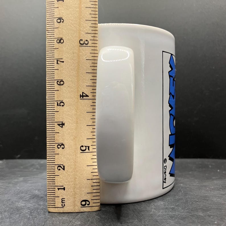 Puede incluir: Taza de cer&aacute;mica blanca con asa curva. La taza tiene un gr&aacute;fico azul y negro que dice "Mickey" y el logotipo de Disney. La taza mide aproximadamente 9 cm de alto.