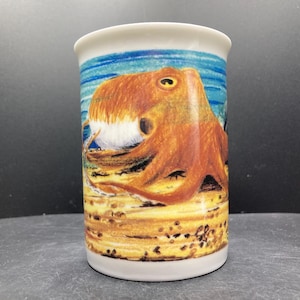 May include: A white ceramic mug featuring a detailed illustration of an orange octopus on a sandy seabed. The background shows a blue sky and ocean. The mug has a cylindrical shape and a smooth, glossy finish.