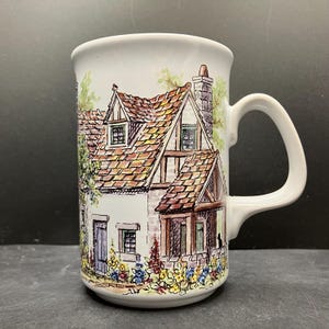 May include: White ceramic mug with a detailed illustration of a cottage. The cottage has a brown tiled roof, dormer windows, and a chimney. The cottage is surrounded by flowers and greenery. The mug has a curved handle.