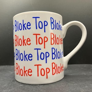 Op de afbeelding: Witte keramische mok met de woorden "Bloke Top Bloke" in blauwe en rode letters. De mok heeft een gebogen handvat en staat tegen een donkere achtergrond.
