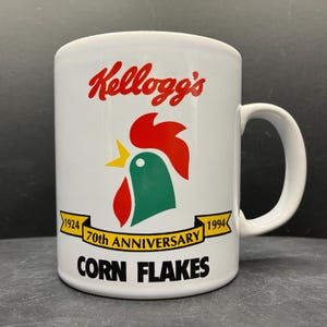 Op de afbeelding: Witte keramische mok met het Kellogg's-logo in rood schrift, een groene en rode haan-afbeelding en de tekst "70th Anniversary Corn Flakes" met de jaren 1924 en 1994. De mok heeft een gebogen handvat.