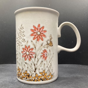 Op de afbeelding: Crèmekleurige keramische mok met een bloemen- en bijenontwerp. De cilindrische mok heeft een gebogen handvat. Het ontwerp bevat oranje bloemen, een bij en bruine grasdetails.