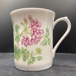 May include: White ceramic mug with a curved handle. The mug features a floral design of pink geraniums and green leaves. The mug is set against a dark background.