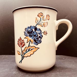 May include: A cream-coloured ceramic mug featuring a dark blue rim and a floral pattern. The design includes a large blue central flower, smaller orange blossoms, and stylised leaves in orange and blue. The mug has a curved handle on the right side.