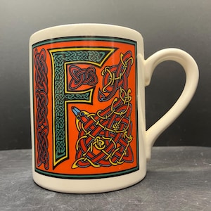 May include: Cream-colored ceramic mug featuring a large, stylized letter "F" in a vibrant orange, green, and yellow Celtic knot design. The mug has a curved handle and a green border at the top. The design is reminiscent of traditional Irish art.