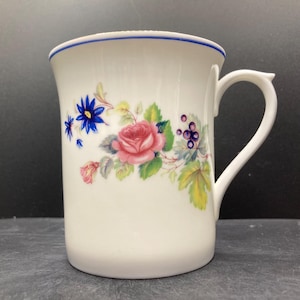 May include: White ceramic mug with a blue rim and handle. The mug is decorated with a floral design featuring pink roses, blue flowers, and green leaves. The mug has a curved handle.