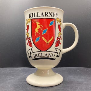 May include: Cream-colored Killarney Ireland mug with a decorative crest featuring a gold deer, blue diamonds, and yellow feathers on a red background. The mug has a pedestal base and a curved handle.