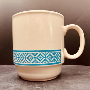May include: A cream-colored ceramic mug featuring a horizontal band of bright blue geometric lattice patterns with small floral accents. The mug has a smooth, cylindrical body with a stepped base and a curved handle on the right side.