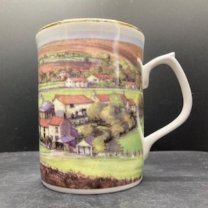 May include: A white ceramic mug depicting a rural landscape with houses, red roofs, and green fields. The mug has a gold rim and a curved handle. The scene is reminiscent of a village.