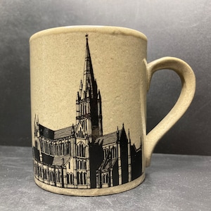 May include: A beige ceramic mug with a black silhouette of a cathedral on the front. The cathedral has a tall spire and intricate details.