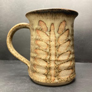 Op de afbeelding: Een bruine keramische mok met een bladerenontwerp. De mok heeft een bruine glazuur met een lichtbruin bladerenontwerp.