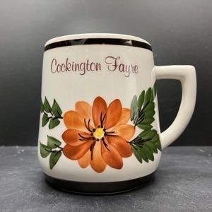 May include: White ceramic mug with a black rim and base, featuring an orange flower design with green leaves. The mug has the text "Cockington Fayre" in brown script. The handle is white.