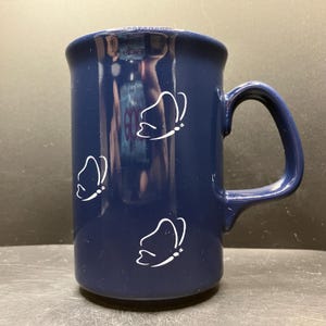 May include: A navy blue ceramic mug with a large handle. The mug features three white butterfly outlines. The mug is cylindrical and has a glossy finish. The mug is suitable for hot or cold beverages.