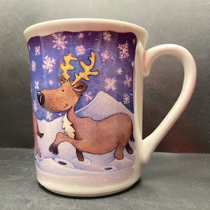 Op de afbeelding: Witte keramische mok met een feestelijke winterscène. De mok heeft een cartoon rendier met gouden geweien, tegen een blauwe achtergrond met sneeuwvlokken en een berg. De mok heeft een gebogen handvat en een witte rand.