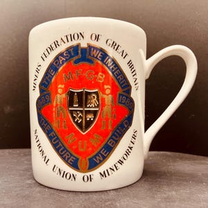 Könnte beinhalten: Ein weißer Keramikbecher mit einem Gedenkemblem der Miners Federation of Great Britain und der National Union of Mineworkers. Das Design enthält den Text The Past We Inherit, The Future We Build, 1889, 1989 sowie die Abkürzung M.F.G.B.