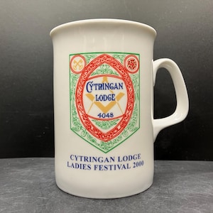 May include: White ceramic mug with a handle, featuring a colorful emblem with the words "CYTRINGAN LODGE" and "LADIES FESTIVAL 2000". The emblem includes a square and compass symbol, with red, green, and gold accents.