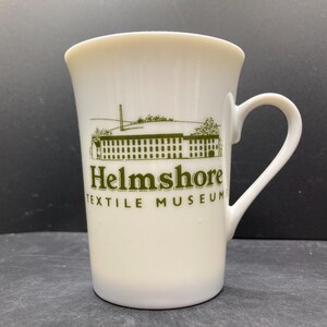 May include: Cream-colored ceramic mug with a curved handle. The mug features a green illustration of a building and the text "Helmshore TEXTILE MUSEUM" in green. The mug has a slightly flared top and a smooth surface.