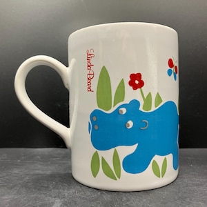 May include: White ceramic mug featuring a blue hippo illustration with green grass and red flowers. The mug has a curved handle and the text "Linda Beard" in red. The design is whimsical and colorful.