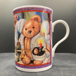 May include: A white ceramic mug featuring a cartoon teddy bear in a striped robe and nightcap, holding a candle and a mirror. The mug has a purple border and a white handle. The mug is approximately 10 cm tall.