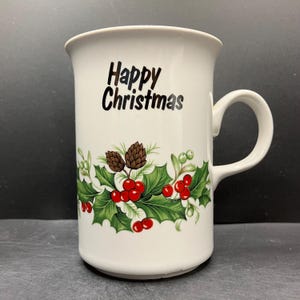 May include: White ceramic mug with a curved handle, featuring the text "Happy Christmas" in black. The mug is decorated with a festive design of holly leaves, red berries, and pinecones, perfect for holiday beverages.