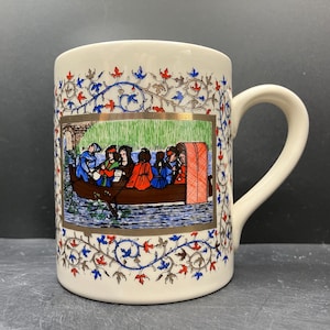 May include: Cream-colored ceramic mug with a decorative medieval scene. The mug features a detailed illustration of people in a boat, framed by a gold border. The design is surrounded by a pattern of red, blue, and gold floral elements. The handle is a matching cream color.