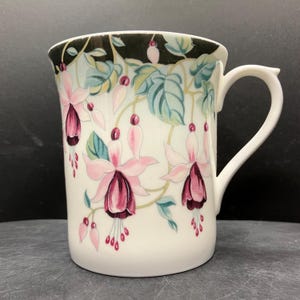 May include: White ceramic mug with a floral pattern of pink and burgundy fuchsia flowers, green leaves, and a black rim. The mug has a curved handle and a cylindrical shape, ideal for tea or coffee.