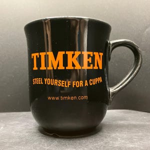 Op de afbeelding: Zwarte keramische mok met het woord "TIMKEN" in oranje blokletters. Daaronder staat de zin "STEEL YOURSELF FOR A CUPPA" en het websiteadres www.timken.com, eveneens in oranje.