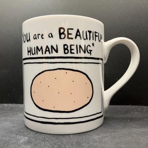 May include: White ceramic mug with a black handle. The mug features the text "You are a BEAUTIFUL HUMAN BEING" above a cartoon potato illustration. The potato is light pink with black dots, and the design is outlined in black.