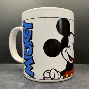 Puede incluir: Taza de cer&aacute;mica blanca con Mickey Mouse. La taza tiene un gr&aacute;fico de Mickey Mouse negro y rojo en la parte delantera, con la palabra "MICKEY" en texto vertical azul. La taza tiene un asa blanca y una l&iacute;nea negra alrededor de la parte superior.
