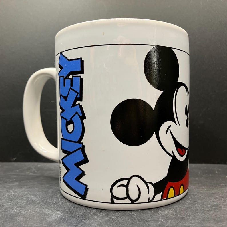 Puede incluir: Taza de cer&aacute;mica blanca con Mickey Mouse. La taza tiene un gr&aacute;fico de Mickey Mouse negro y rojo en la parte delantera, con la palabra "MICKEY" en texto vertical azul. La taza tiene un asa blanca y una l&iacute;nea negra alrededor de la parte superior.