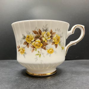 May include: A white teacup featuring a gold-edged base and handle, decorated with yellow rose and green leaf floral motifs. The cup has a ribbed design and a curved handle. Ideal for serving tea or coffee, a classic design.