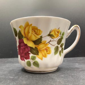 May include: White teacup with a floral design featuring yellow and burgundy roses, green leaves, and gold trim. The cup has a curved handle and a slightly flared rim. A classic design for tea or coffee.
