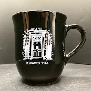 Op de afbeelding: Een glanzend zwarte keramische mok met een witte lijntekening van de hoofdingang van 10 Downing Street. Het ontwerp toont de iconische deur met het nummer 10 en de omliggende ijzeren hekken. De tekst 10 DOWNING STREET staat eronder gedrukt.
