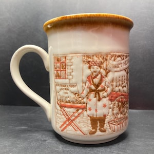 Może przedstawiać: Biały ceramiczny kubek z brązową obwódką. Kubek przedstawia czerwoną i brązową ilustrację ulicznego sprzedawcy, który sprzedaje korzeń Kalibonka, „pewny lek na ból zębów”. Sprzedawca nosi brązową szatę z białymi gwiazdami i brązowy kapelusz. Tekst „Street Herbs” jest napisany na banerze nad sprzedawcą.