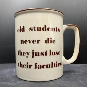 Könnte beinhalten: Cremefarbene Keramik-Tasse mit braunem Rand und Henkel. Die Tasse trägt den Text "old students never die they just lose their faculties" in braunen Buchstaben. Die Tasse hat eine Standardgröße für Kaffee oder Tee.