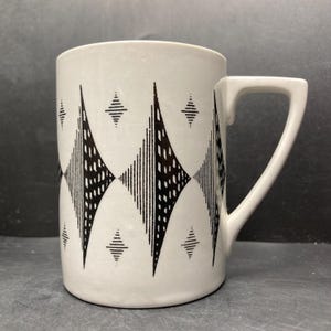 Könnte beinhalten: Weißer Keramikbecher mit einem geometrischen schwarzen Muster. Das Design zeigt Rauten und Linien. Der Becher hat einen quadratischen Griff und eine zylindrische Form. Geeignet für heiße oder kalte Getränke.