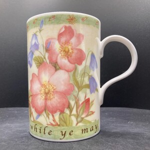 Op de afbeelding: Witte keramische mok met een bloemenontwerp van roze rozen en blauwe klokjes. De mok heeft een gebogen handvat en de woorden "while ye may" staan onderaan gedrukt. De mok is ongeveer 10 cm hoog.