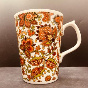 Op de afbeelding: Een hoge keramische mok met een witte basis, versierd met een dicht, herhalend patroon van gestileerde bloemen en bladeren in tinten van gebrand oranje, mosterdgeel, olijfgroen en donkerbruin, voorzien van een effen wit gebogen handvat.