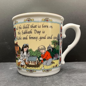May include: A ceramic mug with a white handle and a decorative rim. The mug features a pastoral scene with children and a house, along with the text "And the child that is born on the Sabbath Day is blithe and bonny, good and gay."