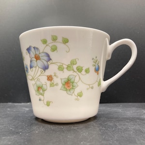 May include: White ceramic teacup with a curved handle, adorned with a floral pattern in shades of blue, green, and orange. The design features stylized flowers and vines, creating a delicate and elegant aesthetic. The cup has a smooth, glossy finish.