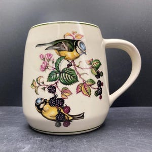 May include: Cream-colored ceramic mug with a green rim and handle. The mug features a detailed illustration of two yellow and blue birds perched on a branch with green leaves, pink flowers, and blackberries. The mug has a curved shape.
