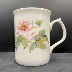 May include: White ceramic mug featuring a pink and yellow floral design with green leaves. The mug has a curved handle and a smooth, glossy finish. The floral pattern is detailed and appears to be hand-painted.